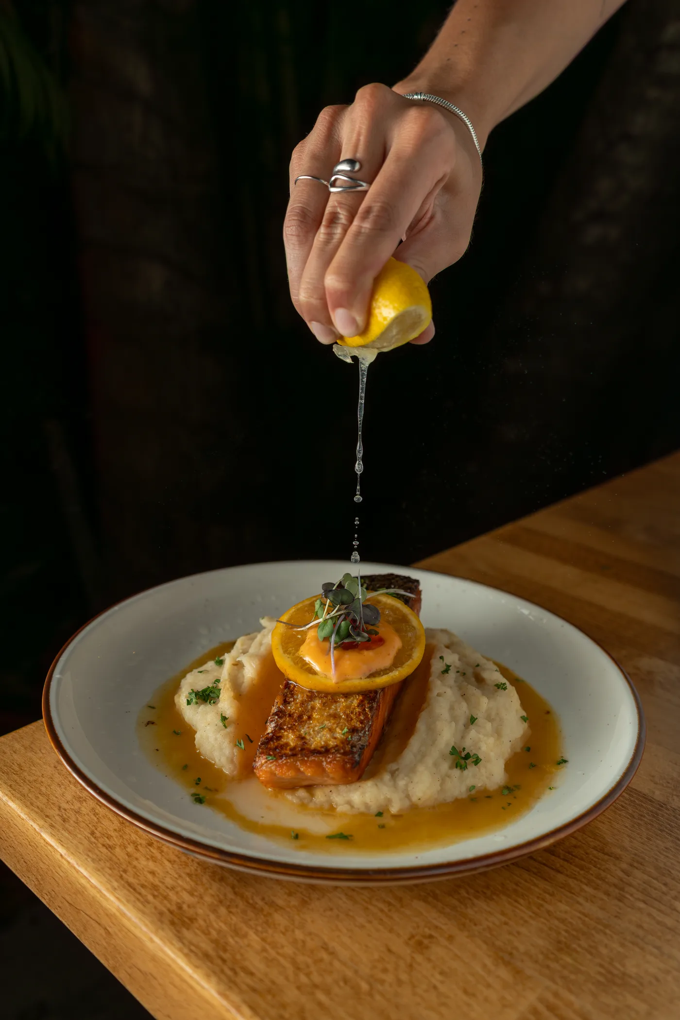 A hand squeezing lemon over crispy salmon and purée