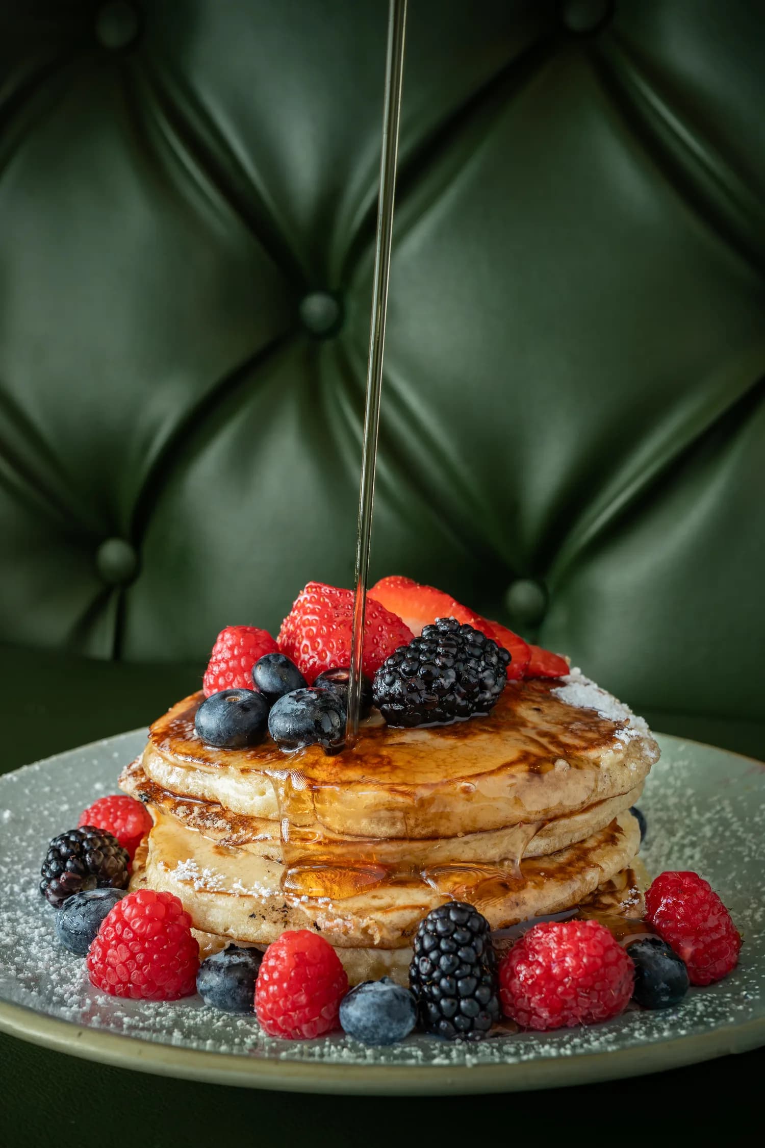 Syrup poured over berry pancakes against a green booth